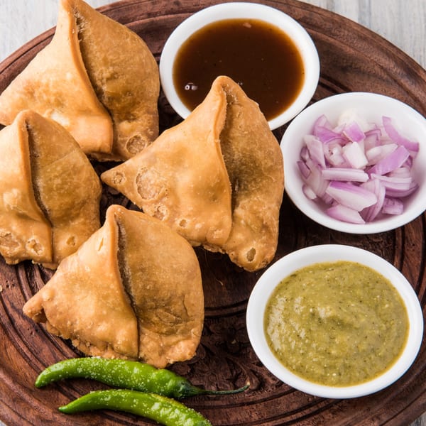Golden-brown Kadai Indian Kitchen samosas with green chili, red onion, and two dipping sauces on a wooden tray.